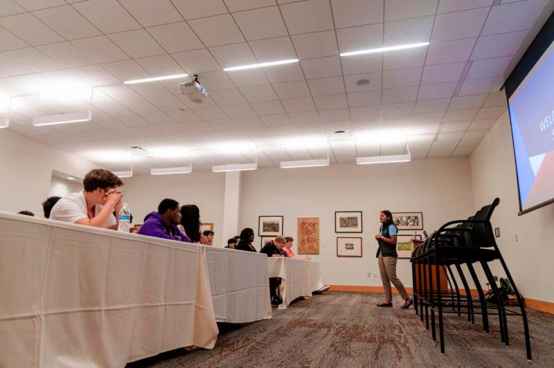 a woman standing in a room with a group of people sitting at tables